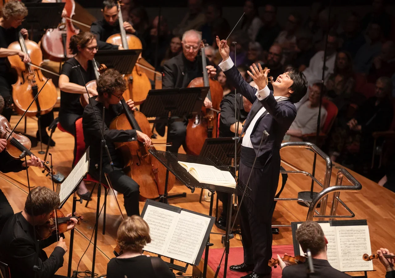 Music Director Kazuki Yamada conducts the City of Birmingham Symphony Orchestra, arms raised, with cellists and a full audience behind him.