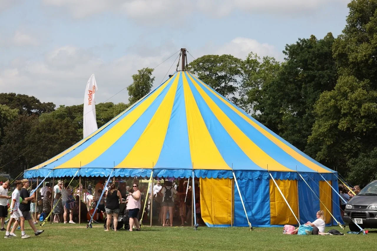 The blue-and-yellow big top of The Great Big Climate Circus stands on the grass under a bright sky, a World Jungle flag flying beside it as visitors gather at the open sides of the tent.