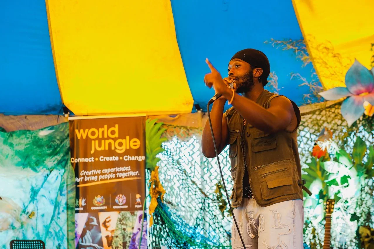 A rap and spoken-word artist performs into a microphone on stage beneath the blue-and-yellow canopy, beside a World Jungle 'Connect · Create · Change' banner and tropical-themed decorations.
