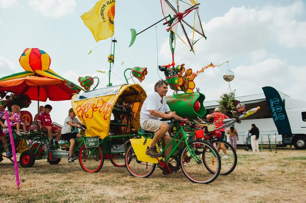 A man pedals the colourful, flower-and-flag-decorated Rinky Dink pedal-powered sound system, with children riding alongside on linked bikes and the Eat Well Wagon community kitchen parked behind.